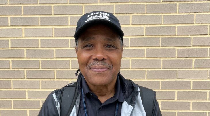 Black man in a MOHS branded hat and jacket smiling while standing in front of a tan brick wall